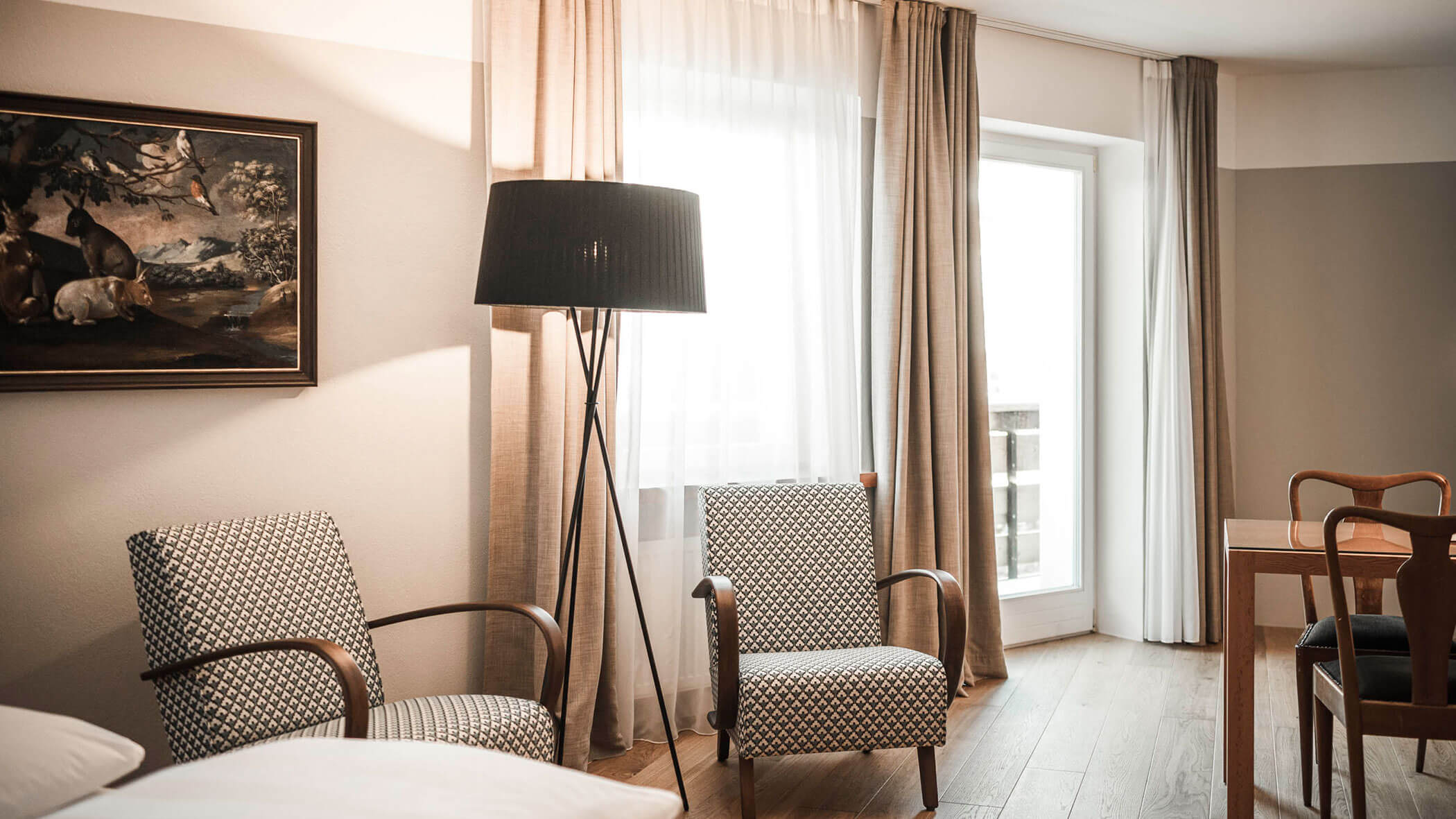 Modern hotel room with two patterned armchairs, a floor lamp, large window, and a framed painting on the wall.