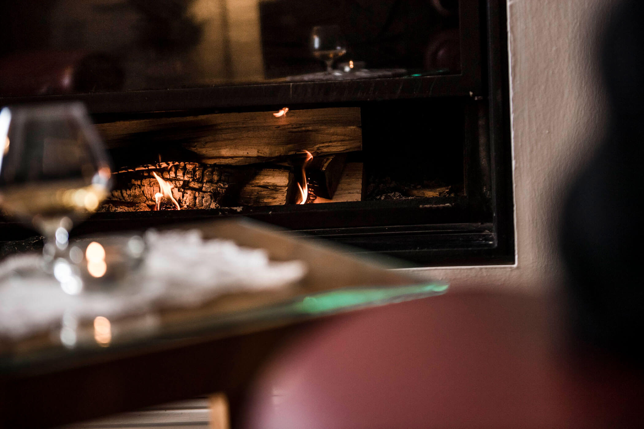 Glass of brandy on table in front of a lit fireplace with burning logs, creating a cozy atmosphere.