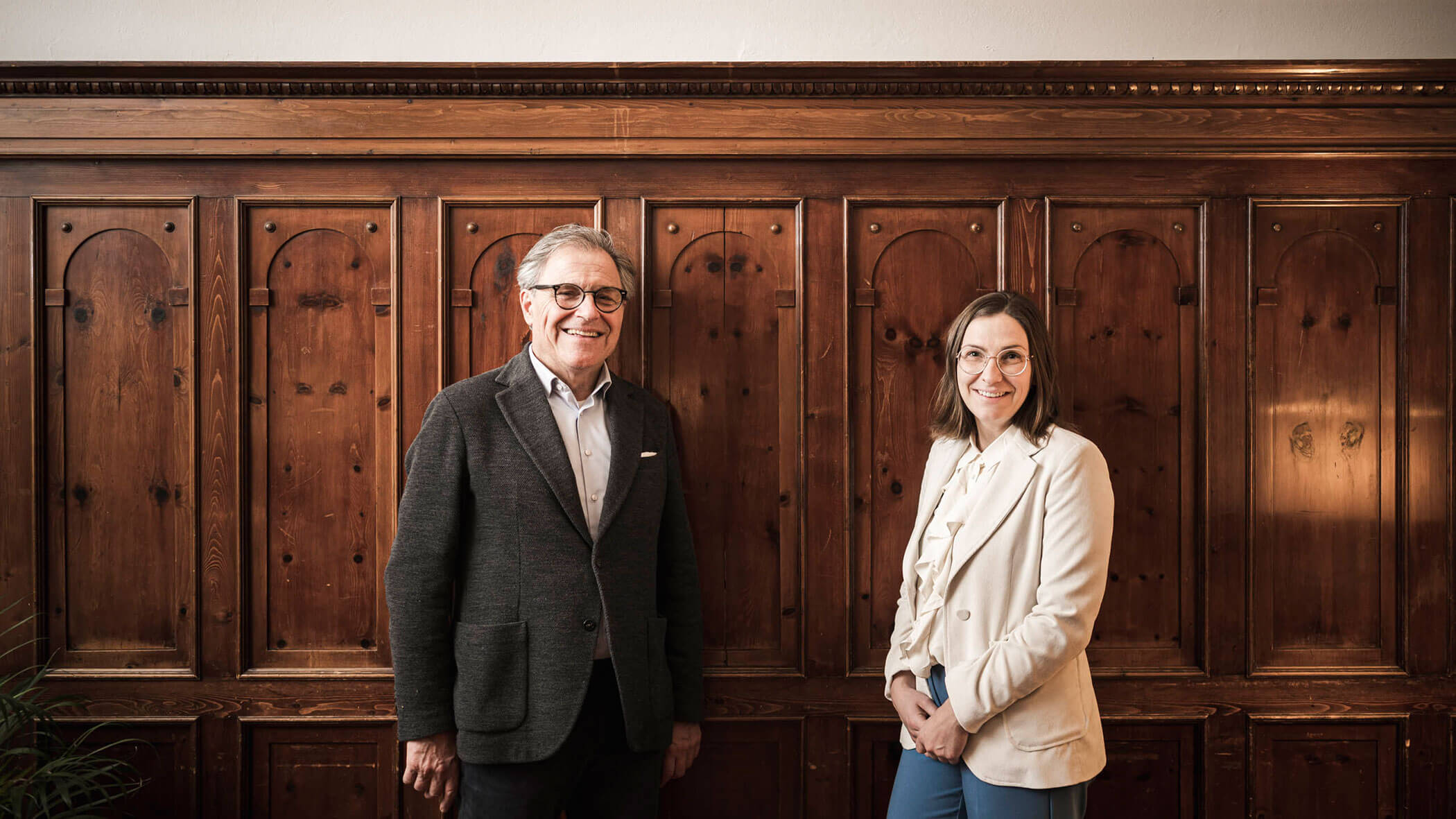 Two people in business attire standing and smiling in front of a wooden paneled wall.
