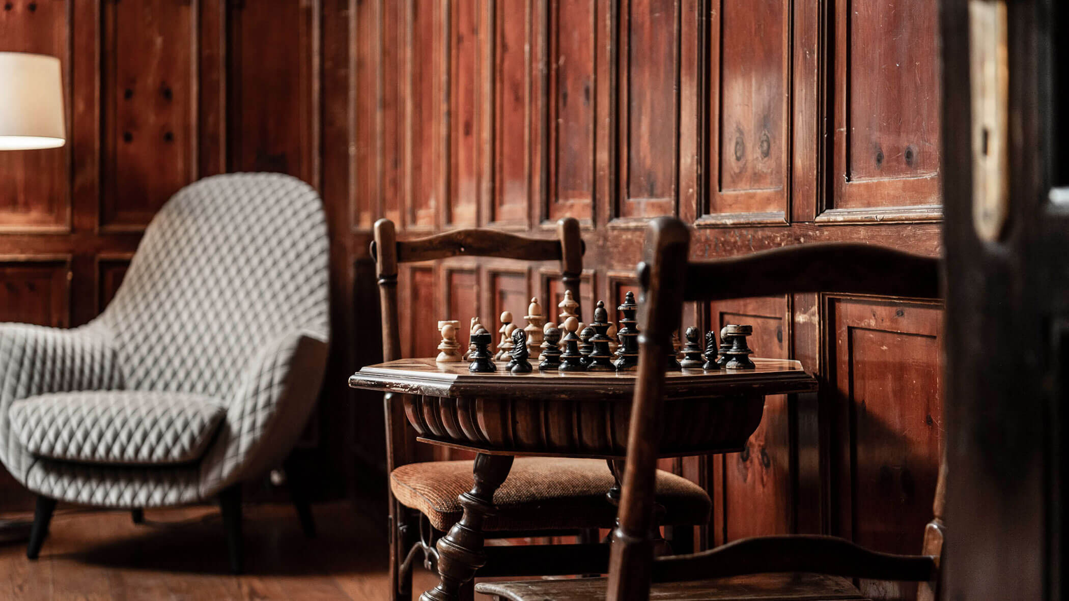 A cozy room with wooden walls, a chessboard on a table, chairs, and a patterned armchair.