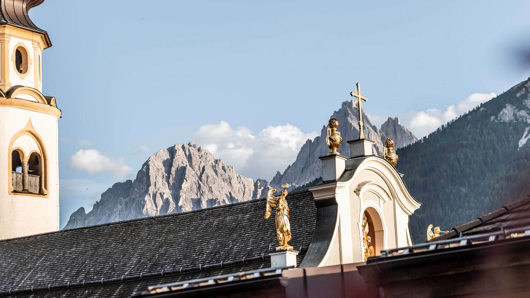 Tetto della chiesa con statue dorate e una croce, sullo sfondo di montagne rocciose e di un cielo parzialmente nuvoloso.