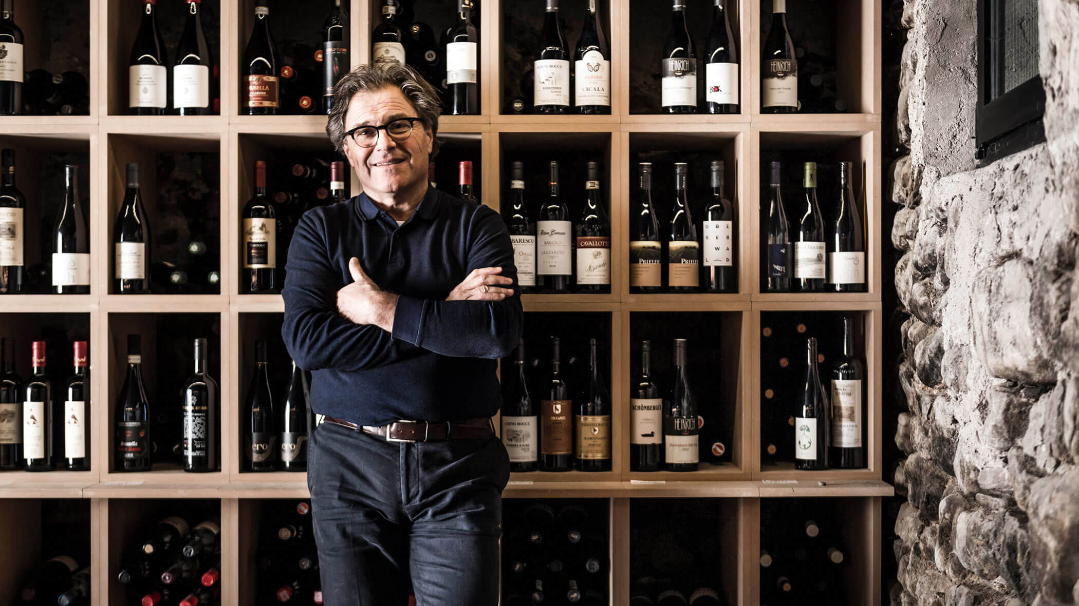Man standing in front of a wine rack filled with bottles, smiling with arms crossed, in a rustic setting.