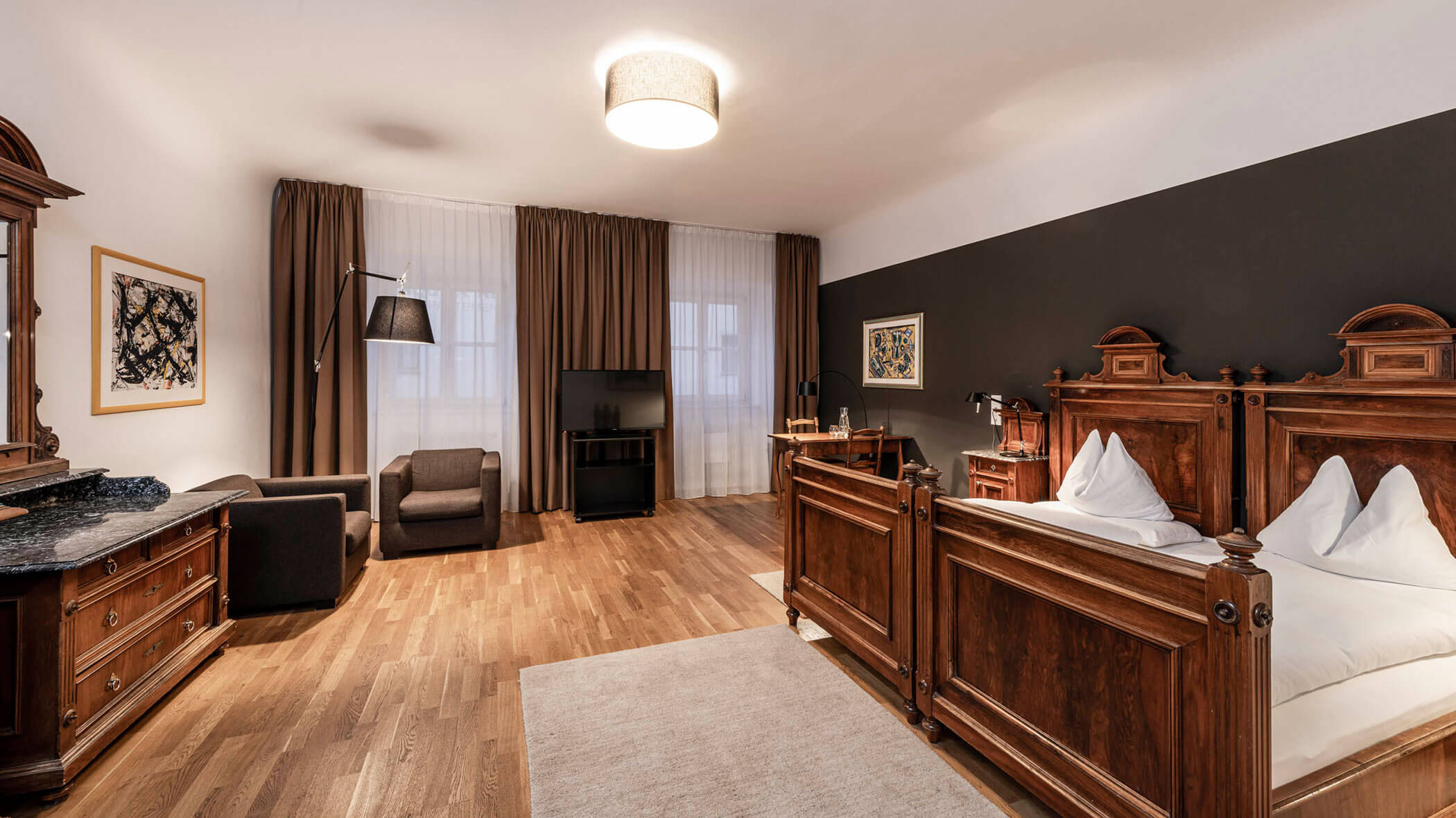 Ampia camera da letto con mobili antichi in legno, letto matrimoniale, poltrona, comò e grandi finestre con tende marroni.