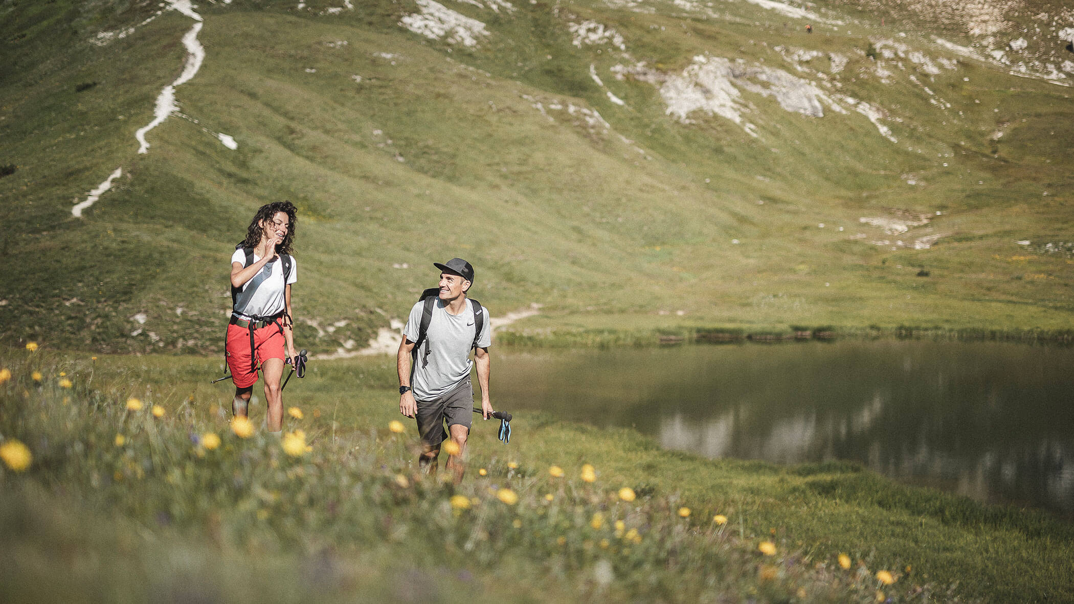 Two people with backpacks hike up a grassy hillside near a lake, surrounded by wildflowers and mountains.