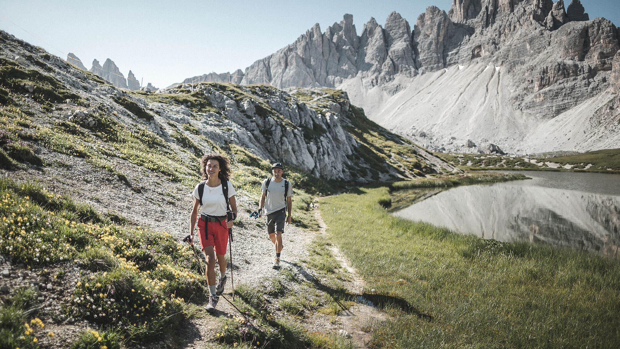 Two people hike on a mountain trail beside a lake, with rocky peaks and green grass under clear skies.