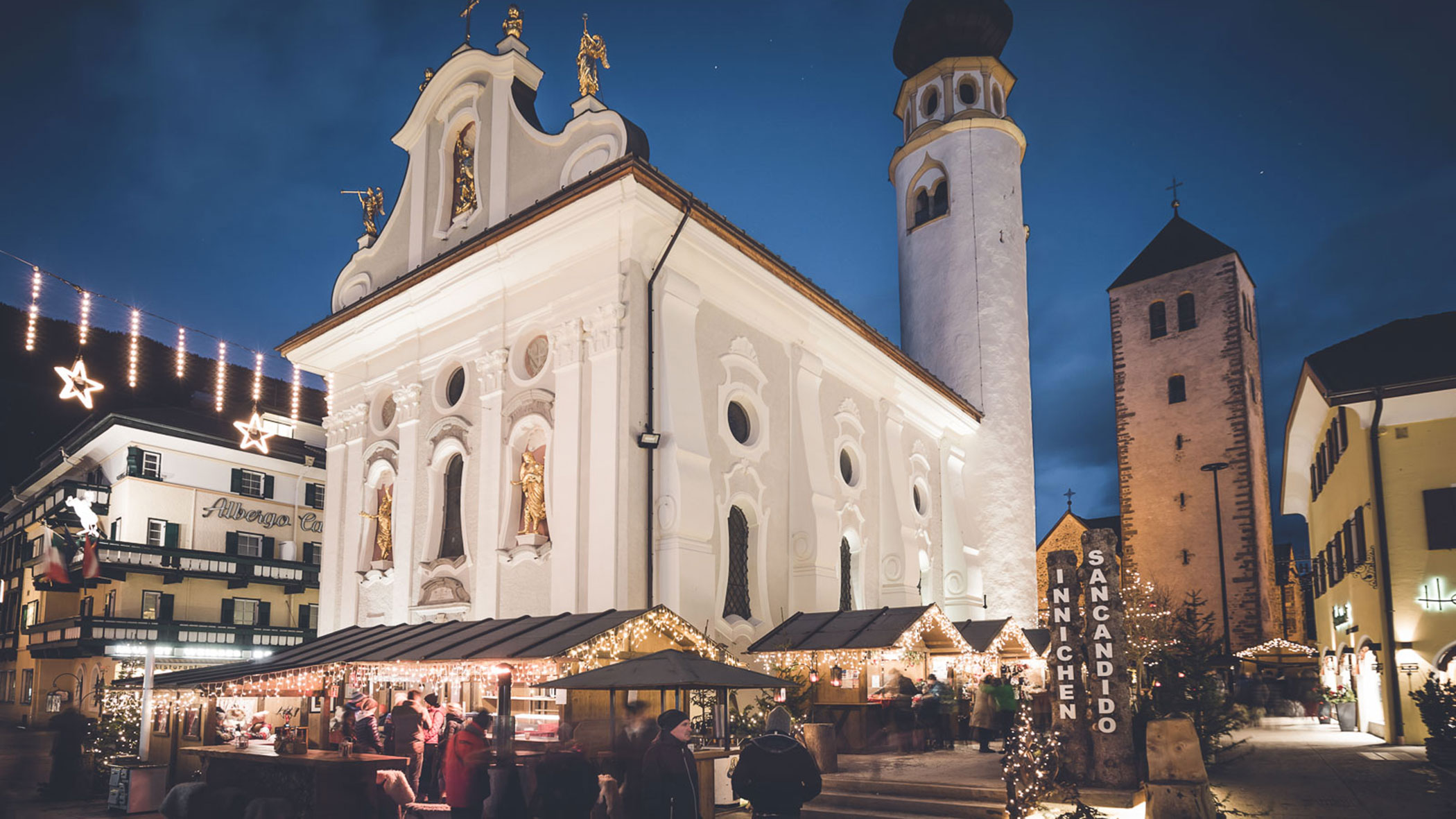 People gather at a festive Christmas market beside a lit-up white church and tower in the evening.