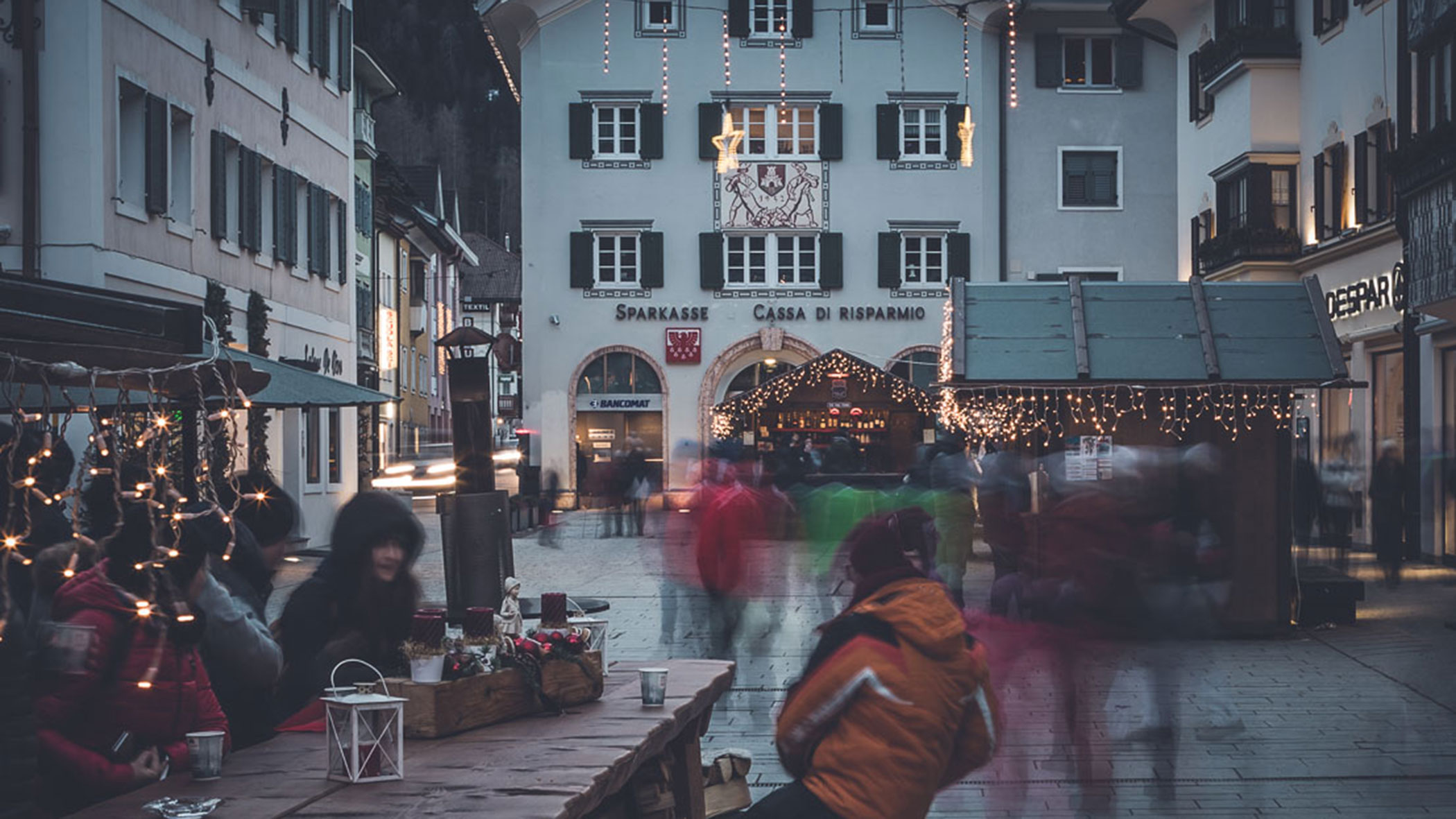 People gather at outdoor tables in a festive, decorated European town square with blurred motion.