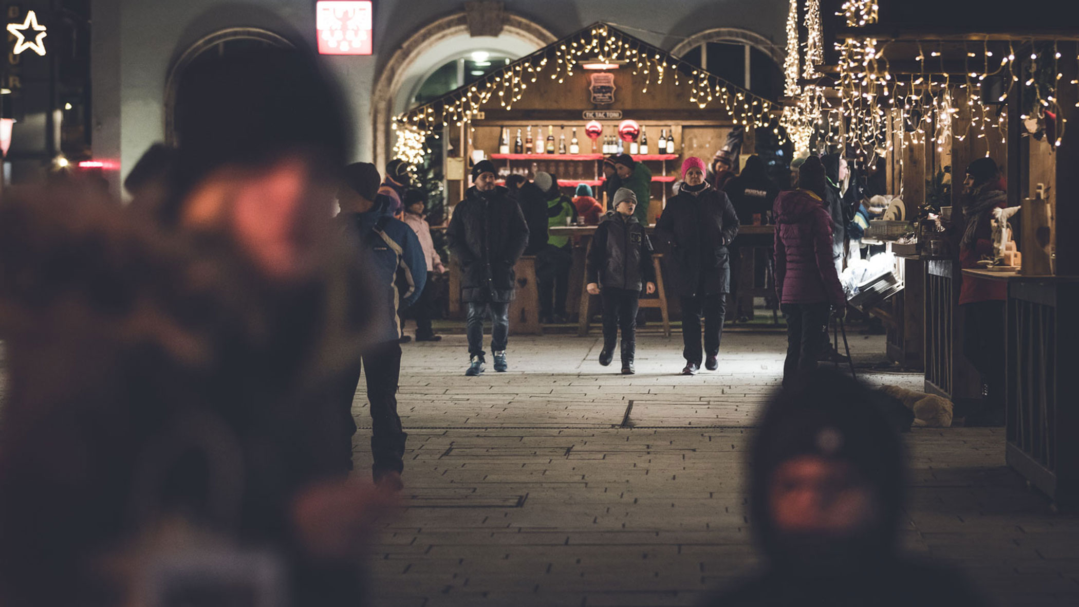 People walk through a festive outdoor market at night, with a wooden booth and string lights in the background.