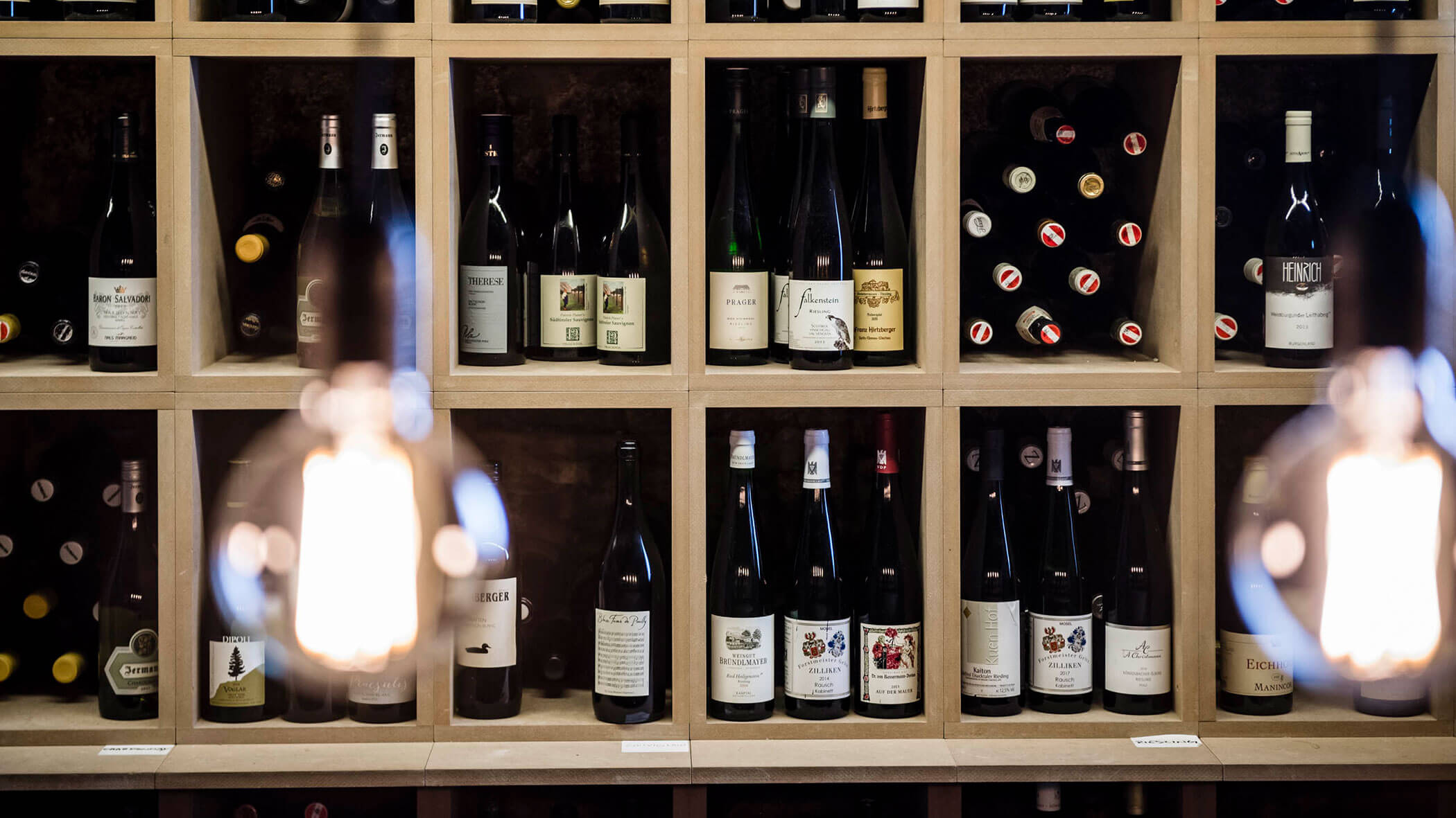 Wooden wine rack filled with assorted wine bottles, with blurred hanging lights in the foreground.