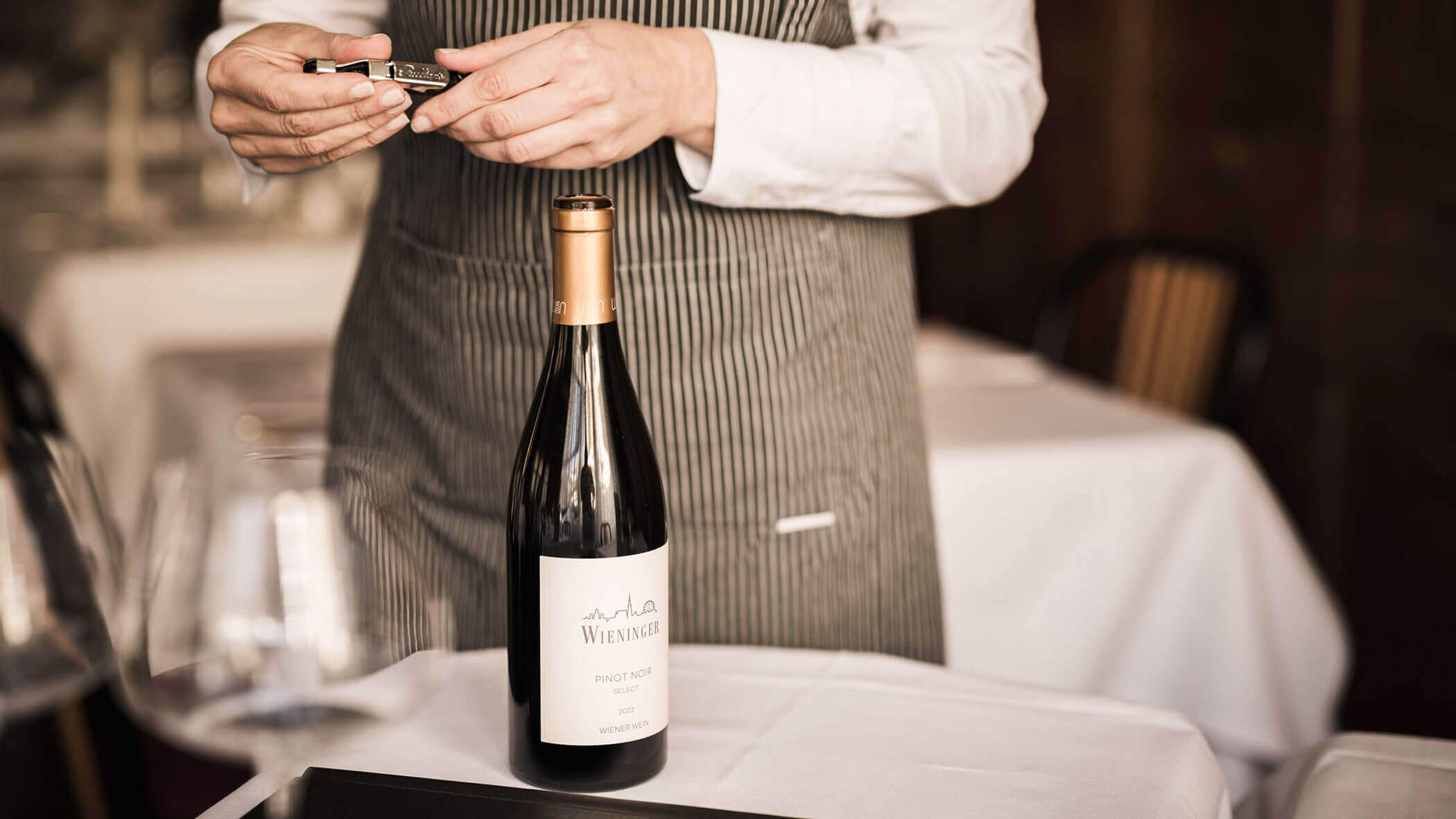 A server in a striped apron opens a bottle of wine at a table set with a white tablecloth.