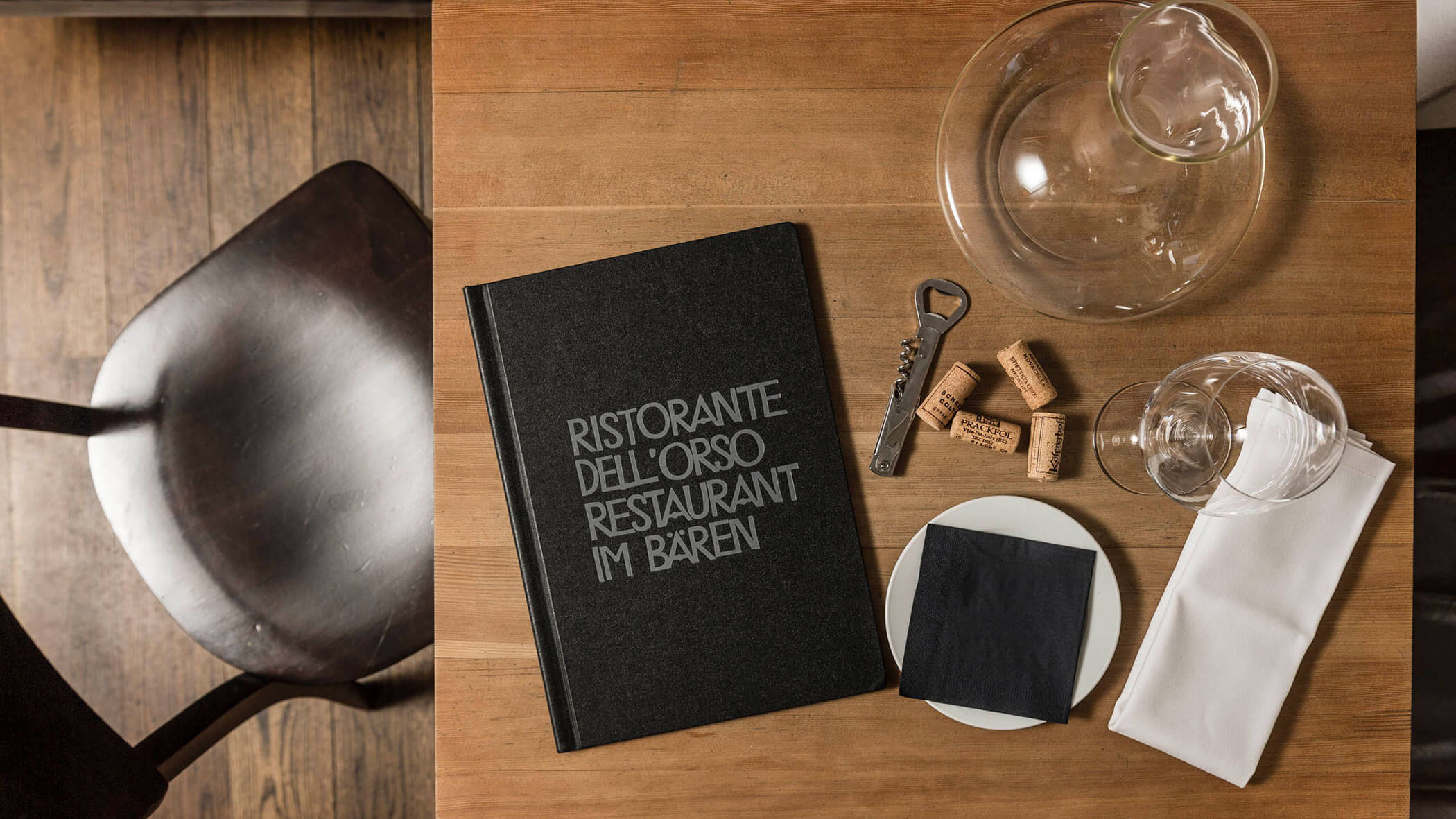 Menu, corks, wineglass, decanter, and napkin on a wooden table next to a dark chair.