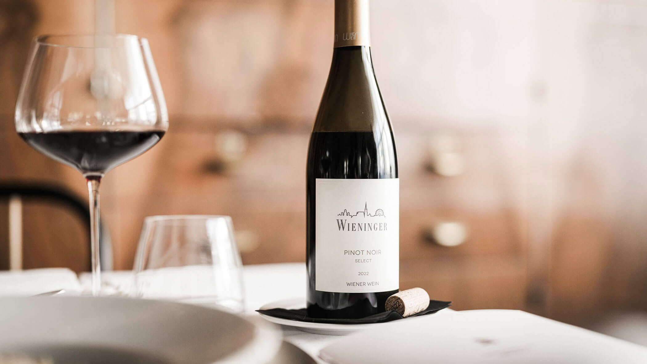 A bottle of Wieninger Pinot Noir wine with a glass of red wine and a cork on a set dining table.
