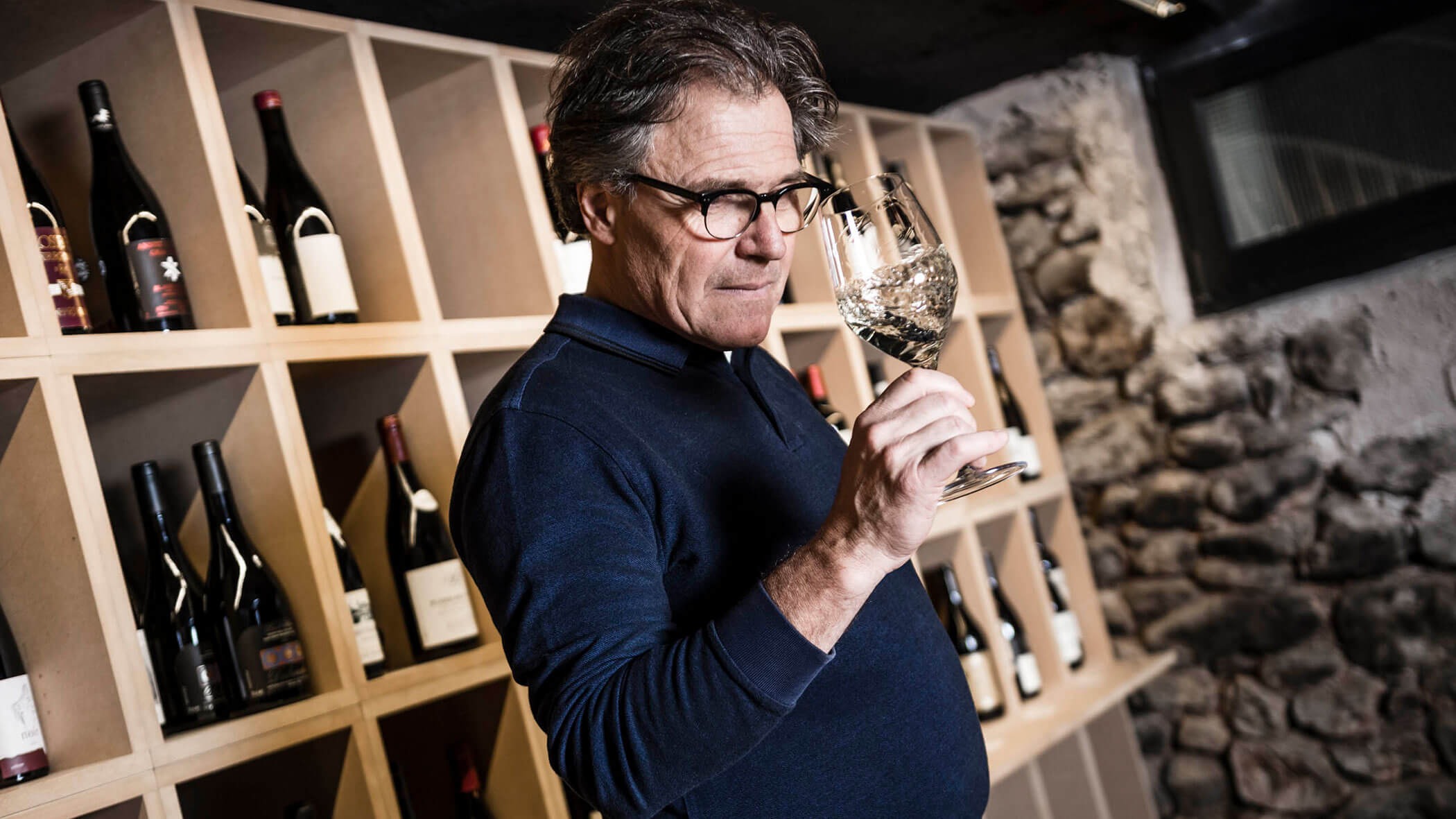 Man in glasses smelling a glass of white wine in a wine cellar with bottles on wooden shelves behind him.