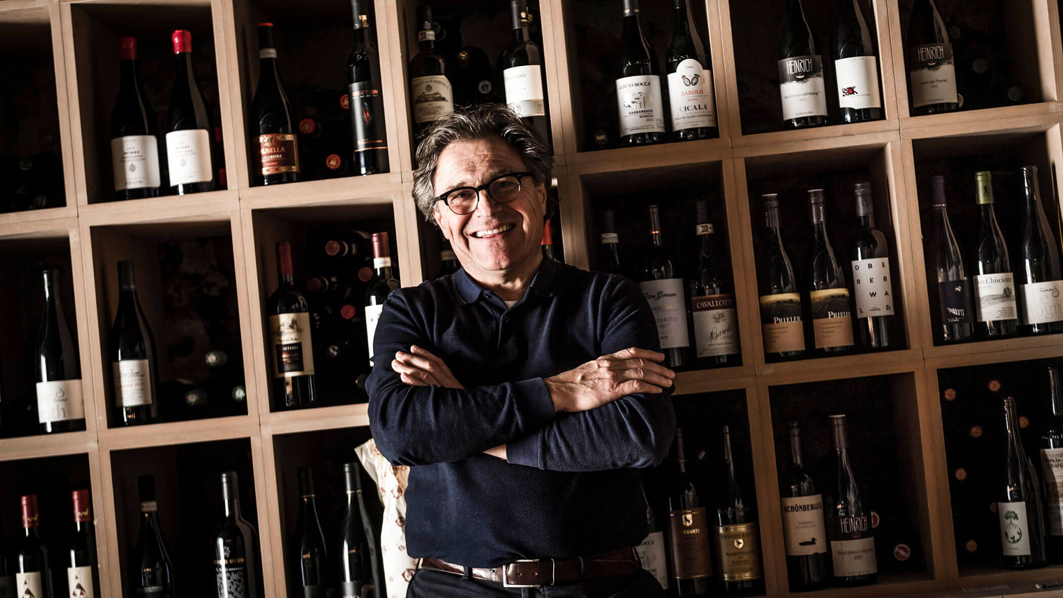 Smiling man with glasses stands in front of shelves filled with wine bottles, arms crossed.