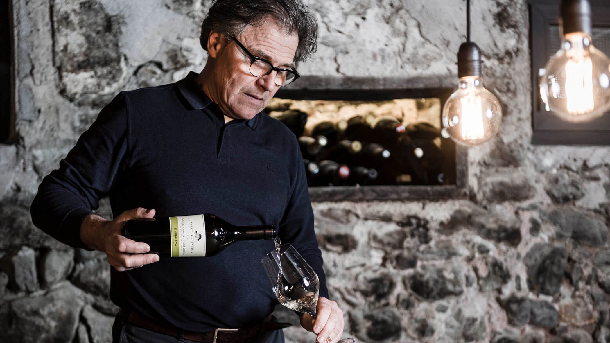 A man pours wine into a glass in a rustic cellar with stone walls and hanging light bulbs.