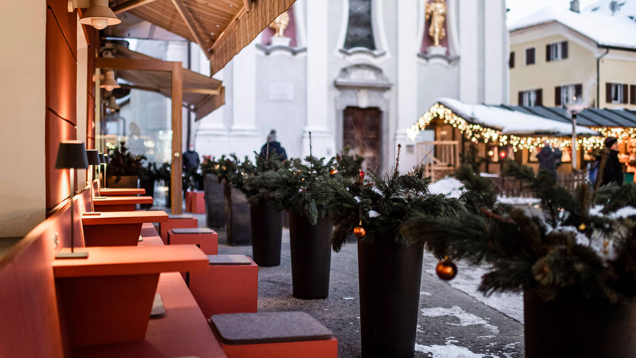 Outdoor cafe with red seating, winter greenery, and holiday lights near a church in a snowy town.
