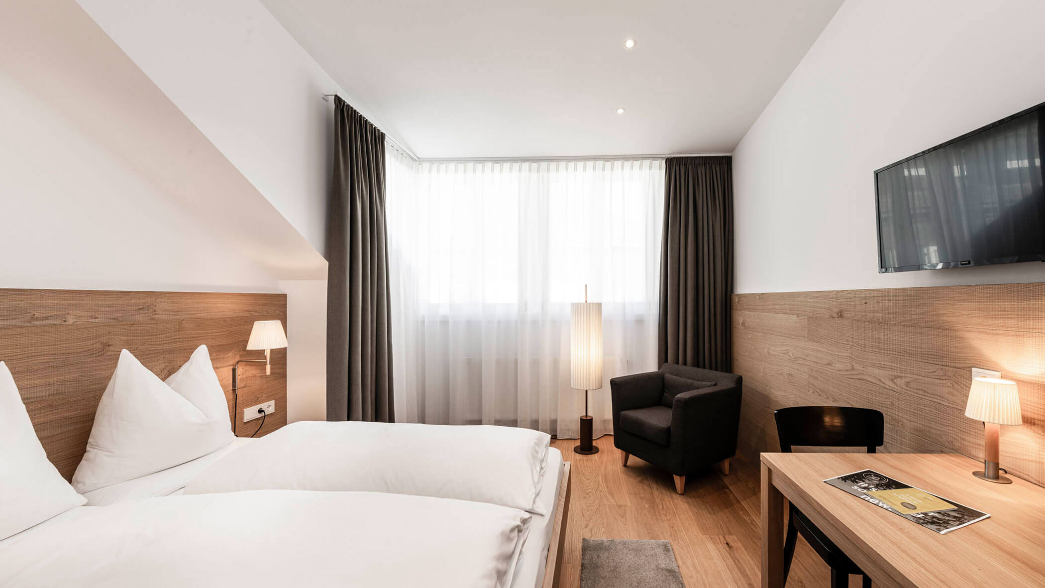 Minimalist hotel room with twin beds, desk, armchair, flat-screen TV, large window, and wooden accents.