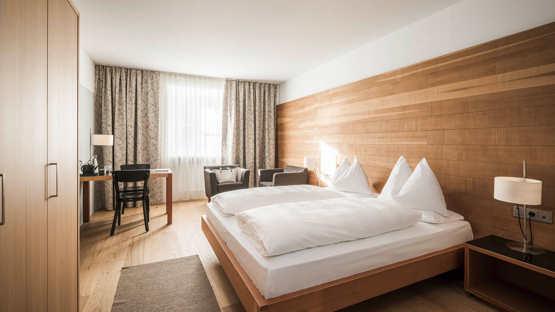 A modern hotel room with a double bed, desk, chairs, large window, and wooden accents.