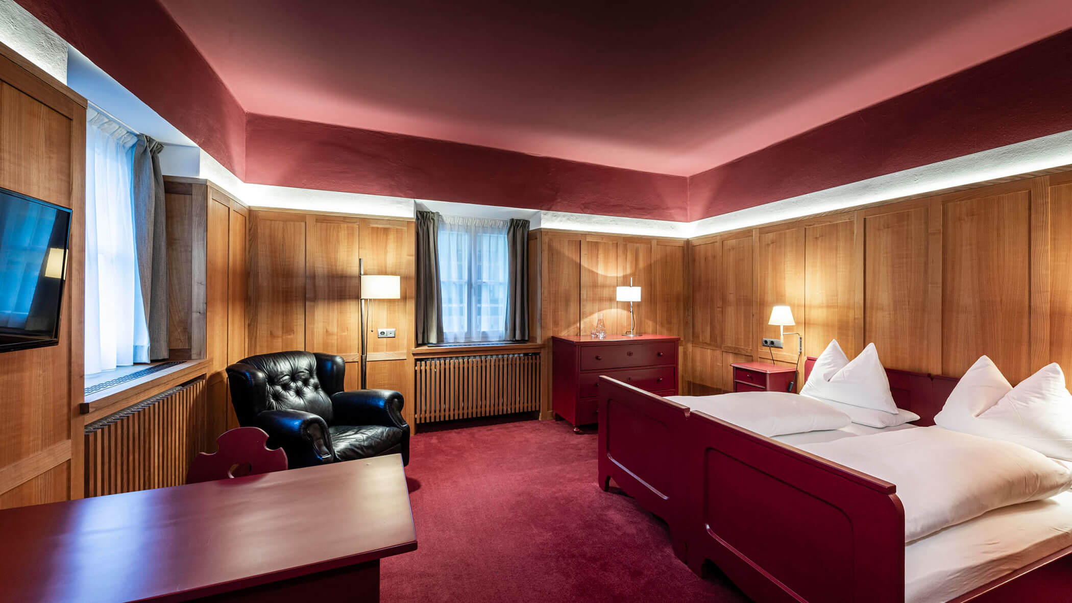 Hotel room with wood-paneled walls, red furniture, a bed, armchair, lamps, and carpeted floor.