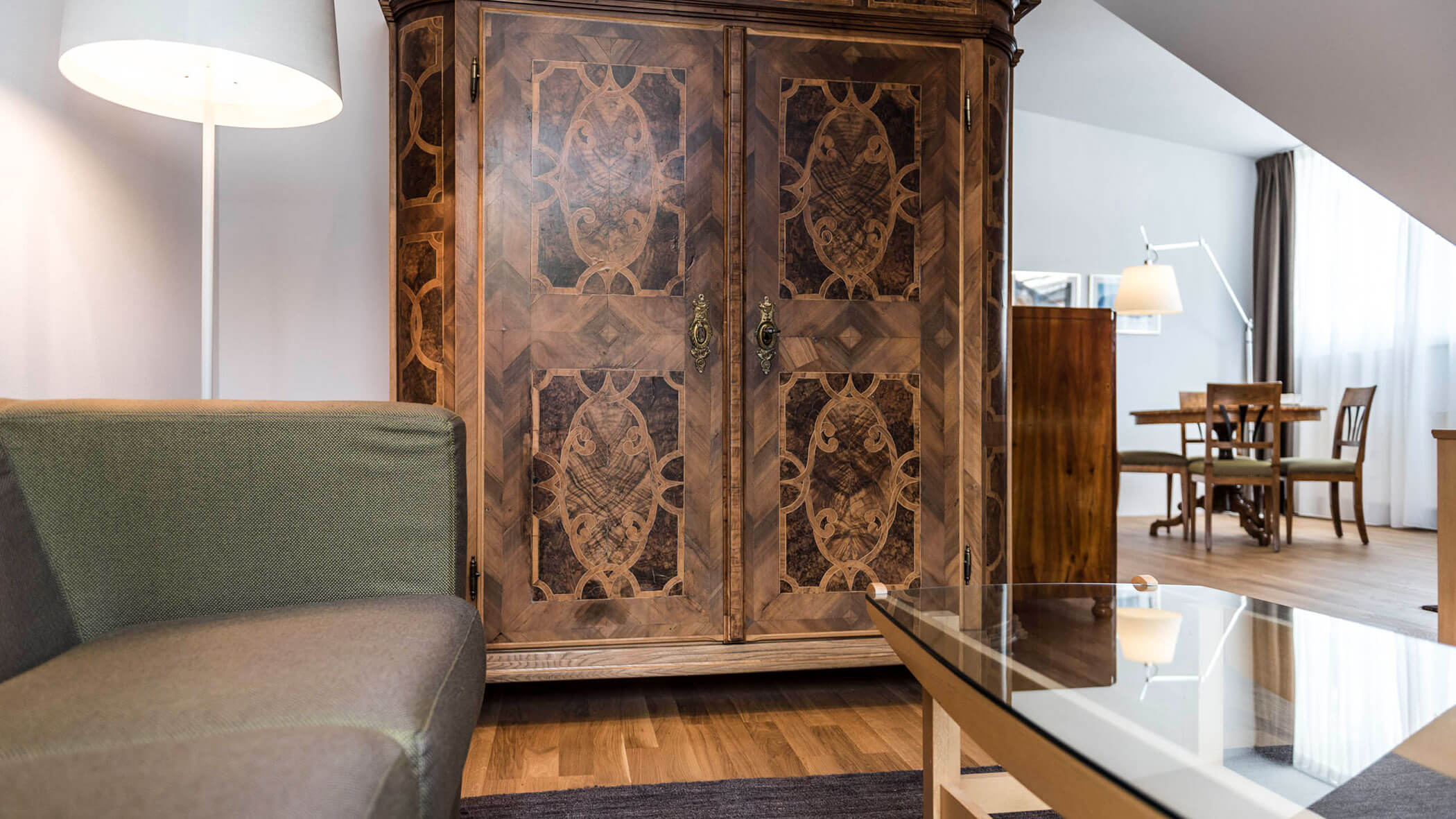 A living room with a patterned wooden armoire, green sofa, glass coffee table, and dining area in the back.