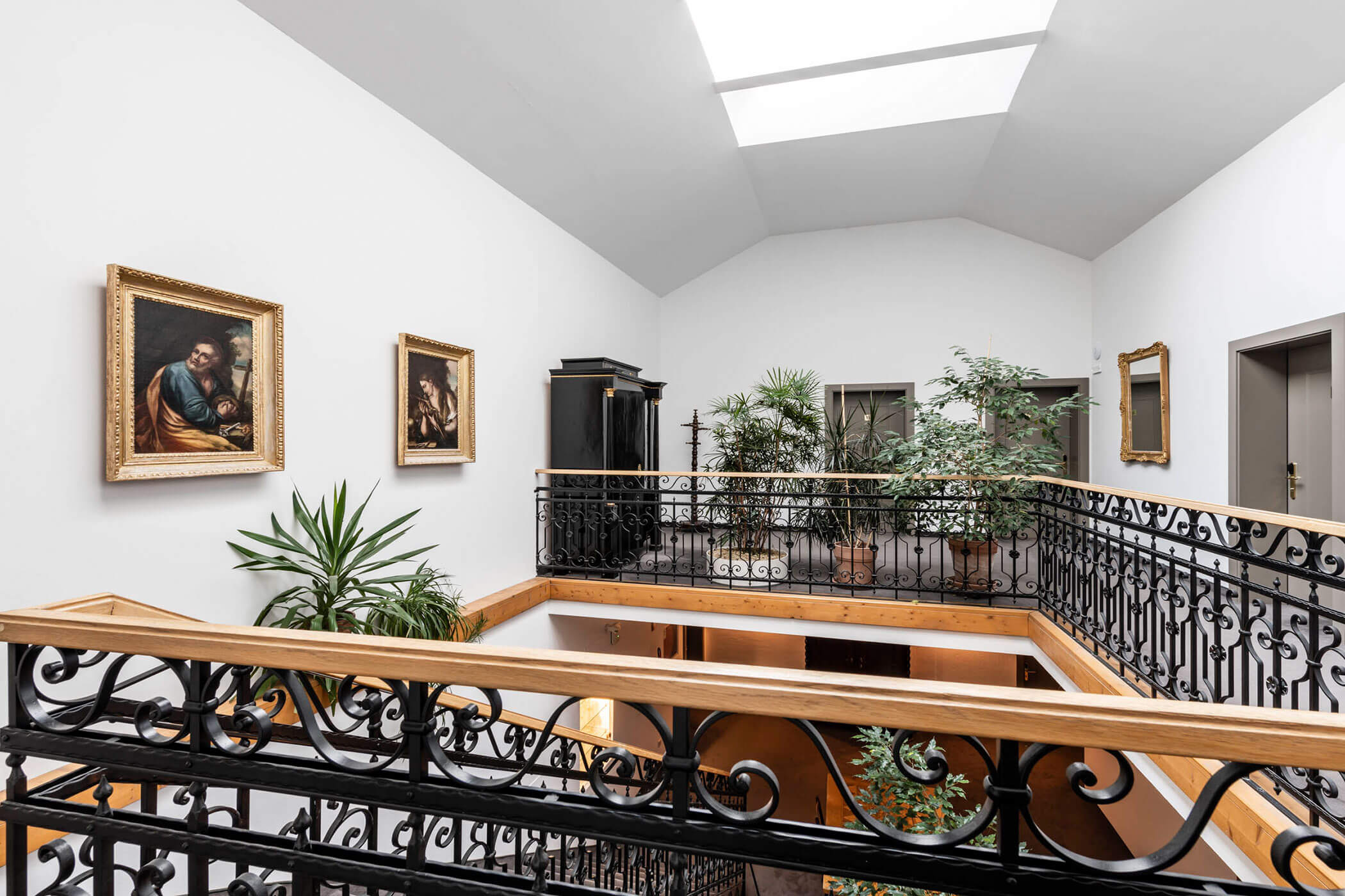 Second-floor hallway with ornate black railings, skylight, paintings, plants, and a black armoire.
